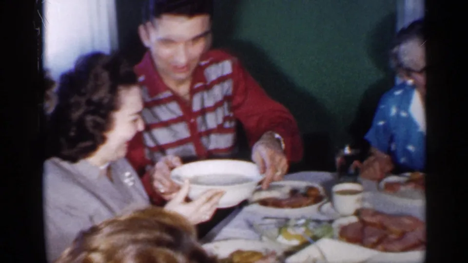 A man in a red and white striped shirt passes a white serving bowl while adults sit around a 1958 New Jersey dinner table.
