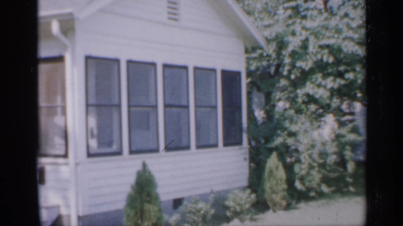 White-sided house with shuttered windows, shrubs, lawn, and trees.