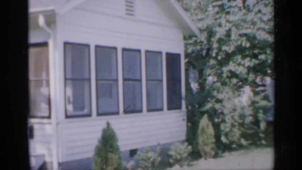 1958 New Jersey home-movie frame showing a white-sided house facade with dark shutters, front shrubs, grass, and leafy trees.