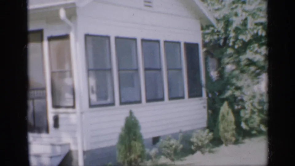 1958 New Jersey frame of a white house exterior with a screened porch entrance, multiple windows, dark shutters, shrubs, and trees.