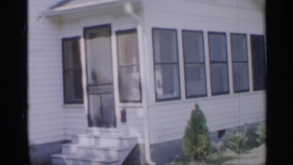 1958 New Jersey home-movie frame centered on a white-sided house entrance with a screen door, concrete steps, windows, shutters, and low planting.
