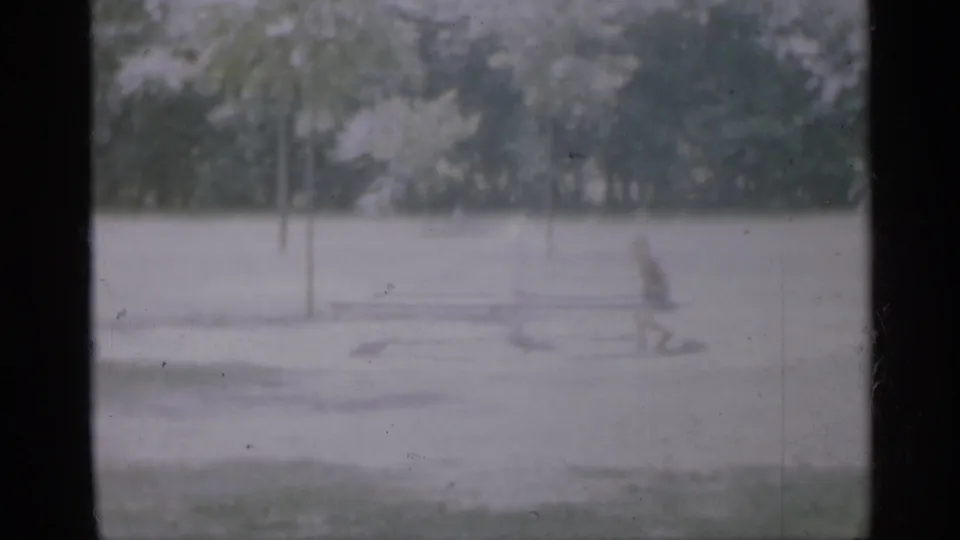 Soft 1958 New Jersey frame showing a small child beside low playground equipment with trees and open pavement behind.