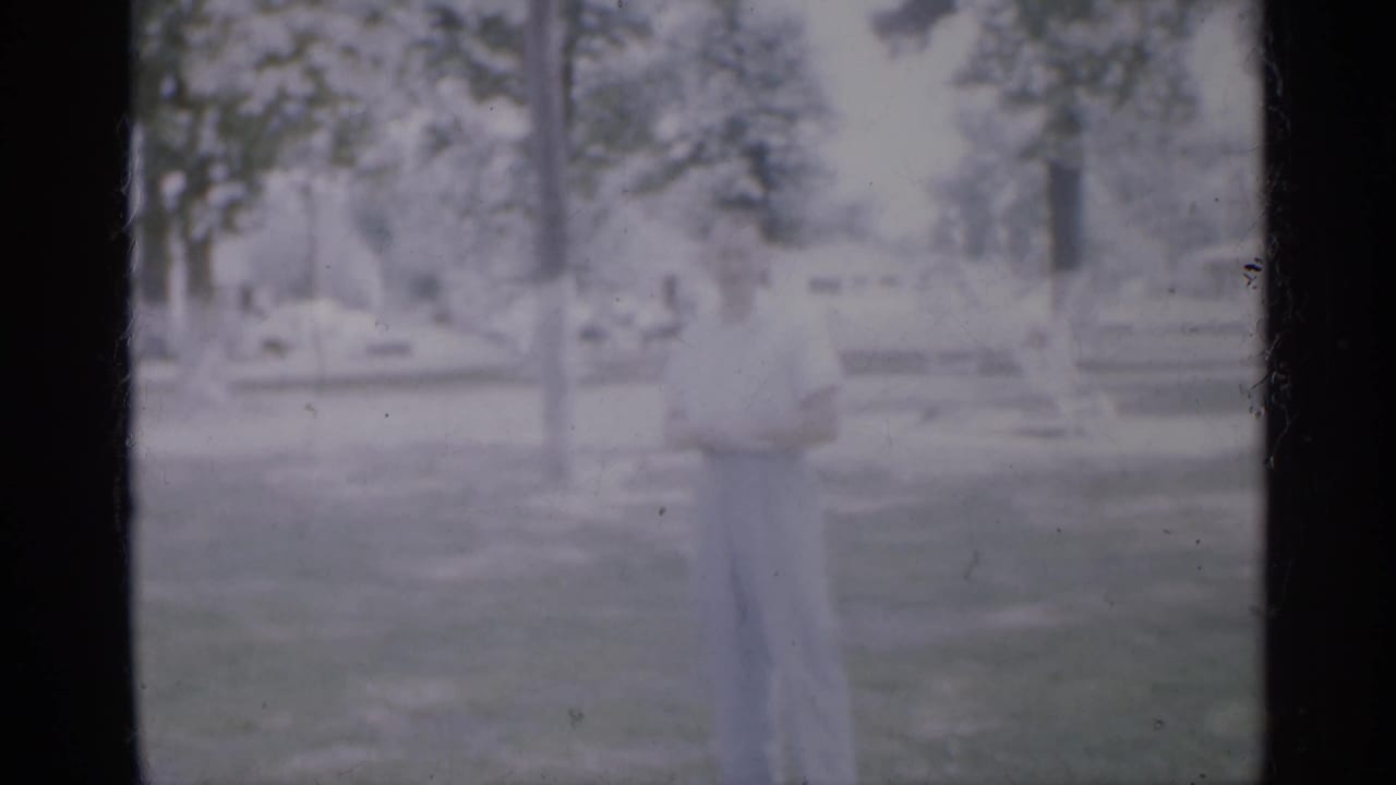 The man remains standing in grass, with trees and low park seating visible behind him in the faded 1958 New Jersey footage.