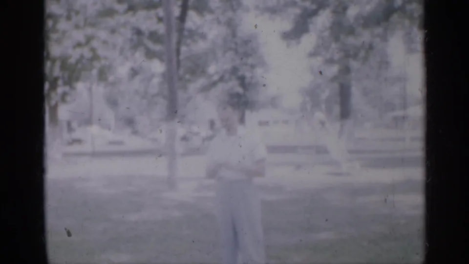 1958 New Jersey home-movie frame showing a small child in light clothing standing in a sunlit park with trees and low park fixtures behind.