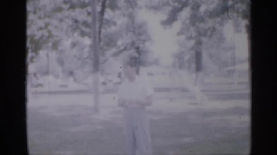 1958 New Jersey park footage with a child in light clothing standing on grass, framed by trees and distant playground or park equipment.