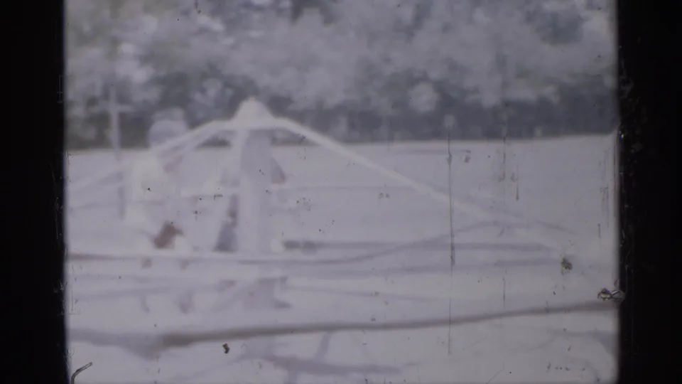 1958 New Jersey home-movie frame showing low playground rails and a small childlike figure in a bright park with trees in the background.