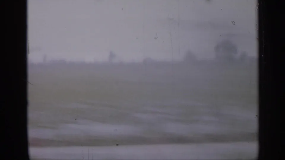1958 New Jersey 8mm frame with a muted open field, distant trees, and dark vehicle-window borders during a moving roadside view.