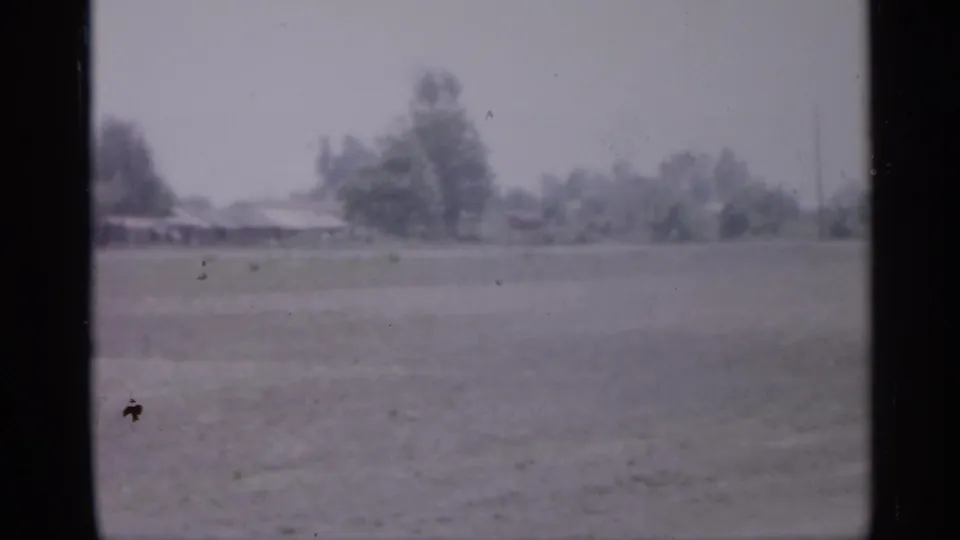 1958 New Jersey 8mm frame showing open ground, distant trees, and a low house or building softened by haze.