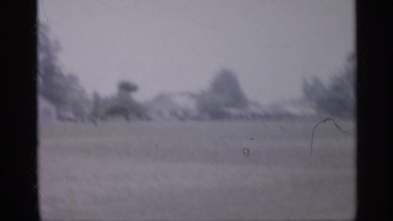 1958 New Jersey home-movie frame showing an open grassy field with distant trees and low houses in soft focus.