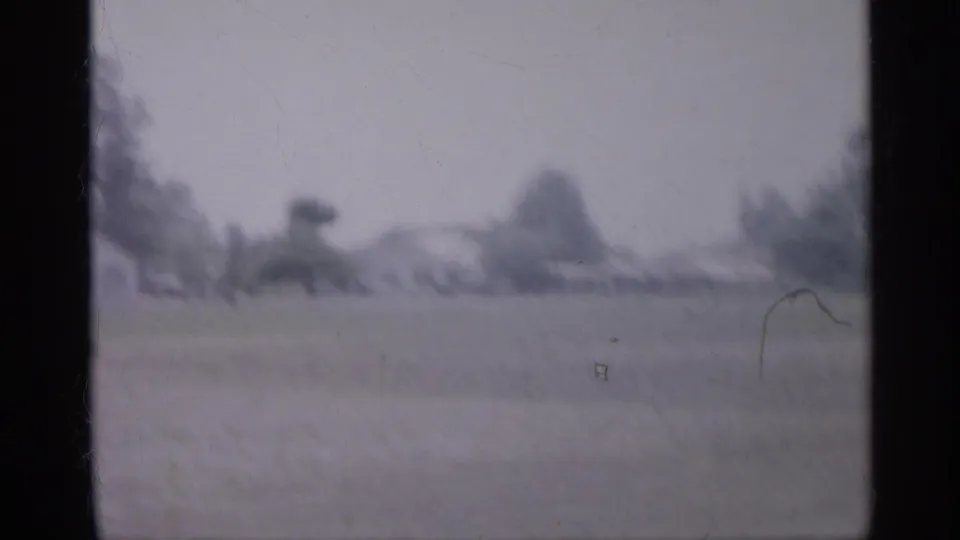 1958 New Jersey home-movie frame showing an open grassy field with distant trees and low houses in soft focus.