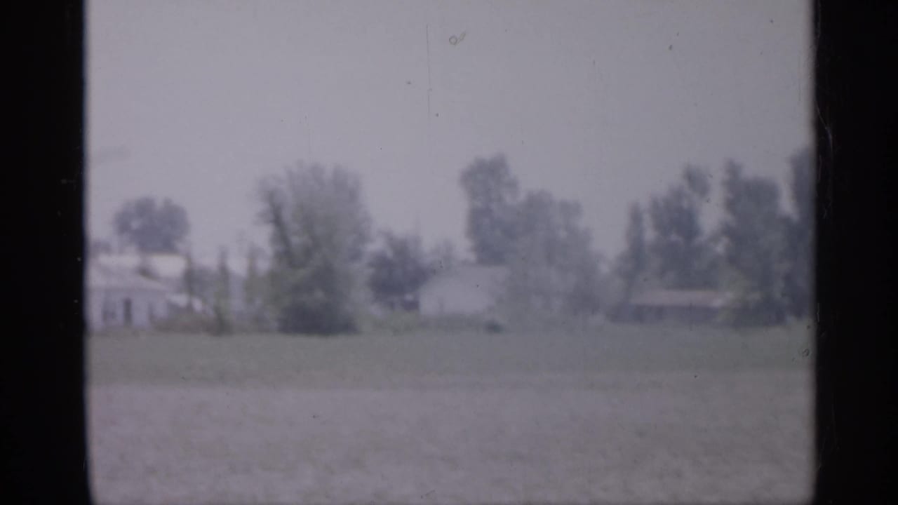 Distant houses and trees beyond a grassy open area, framed by dark film edges.