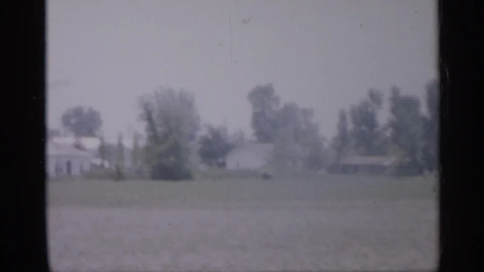 A muted 1958 New Jersey suburban view shows trees and small houses beyond an open field.