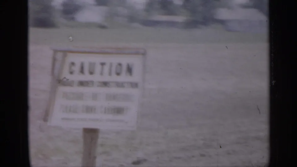 A roadside caution sign stands in a grassy New Jersey suburban setting, with homes and trees blurred behind it.