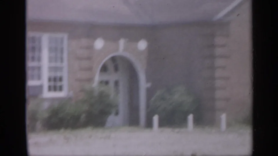 1958 New Jersey 8mm frame showing a brick exterior with an arched doorway, white window, shrubs, and short roadside posts.