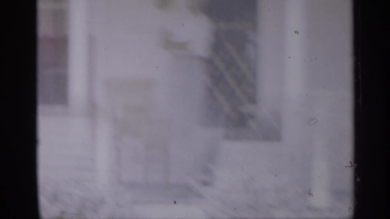 Soft 1958 8mm New Jersey home-movie frame showing a faint man near a doorway or porch area, with dark film borders and exterior structures.