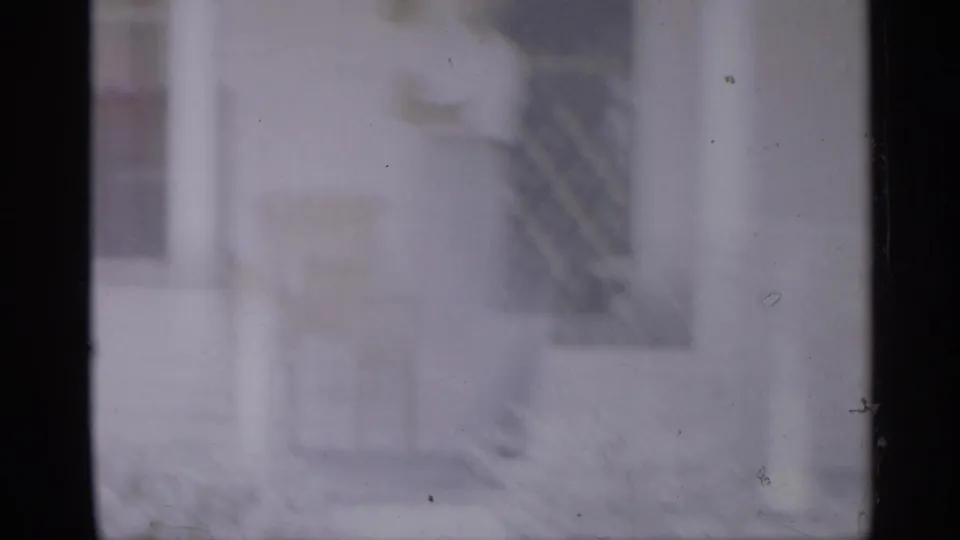Soft 1958 8mm New Jersey home-movie frame showing a faint man near a doorway or porch area, with dark film borders and exterior structures.