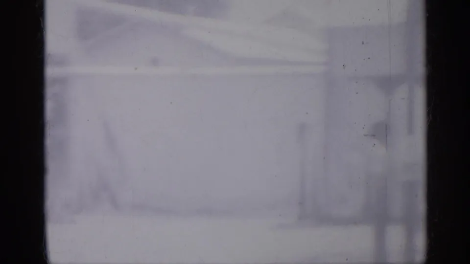 Soft 1958 New Jersey home-movie frame showing a pale tank-like cylinder, fencing, and vertical exterior lines in a residential yard or porch area.