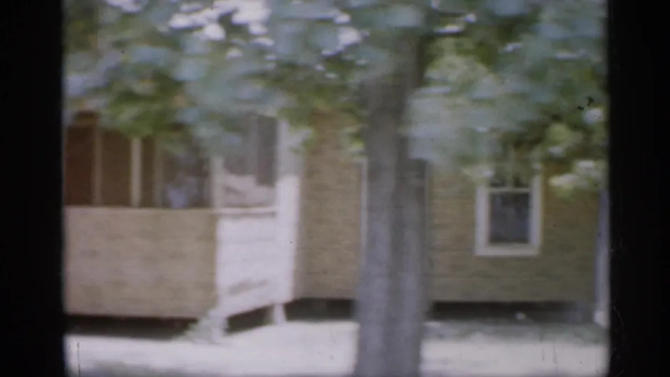 1958 New Jersey home-movie view of a brown house facade, porch rail, window, tree trunks, and grass.