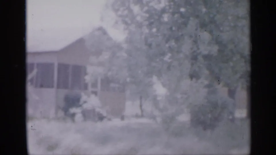 Faded 8mm frame of a New Jersey house facade in 1958, partly hidden by trees, bushes, and a light-colored yard.