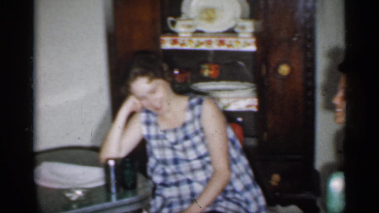 Woman in a blue checked dress seated beside a table in a living room.