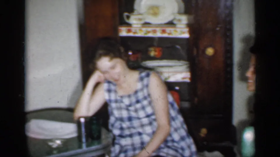 1958 New Jersey home-movie frame showing a woman in a blue checked dress seated near a table, with a dish hutch behind her.