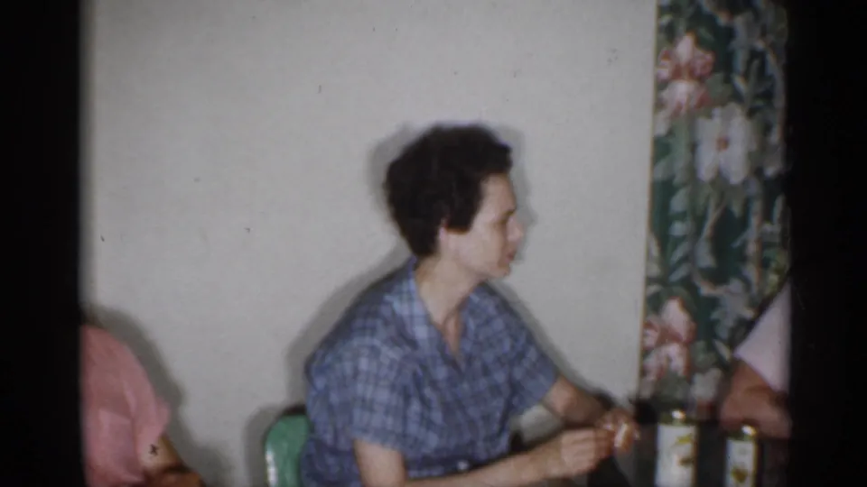1958 New Jersey interior frame with an adult in a blue plaid shirt seated at a table beside floral wall covering and glass bottles.