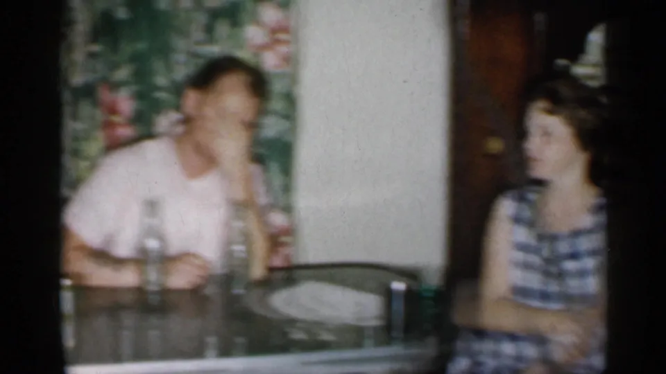 Soft 8mm frame from a 1958 New Jersey home gathering, showing adults seated at a table with bottles, glasses, and floral decor.