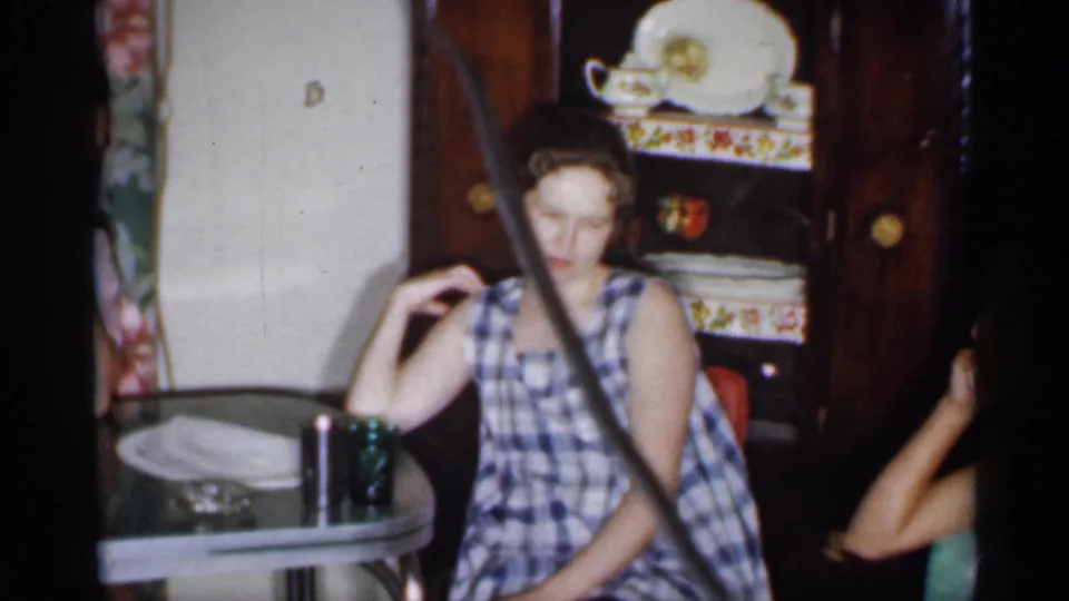 1958 New Jersey home-movie frame showing a woman in a plaid dress seated by a table, with a china cabinet and dishes behind her.