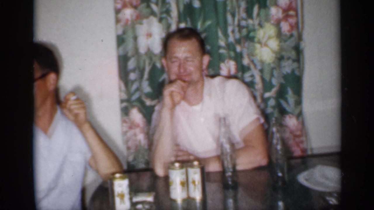 1958 New Jersey home-movie frame showing adult men at a glass table with canned drinks and floral curtains behind them.