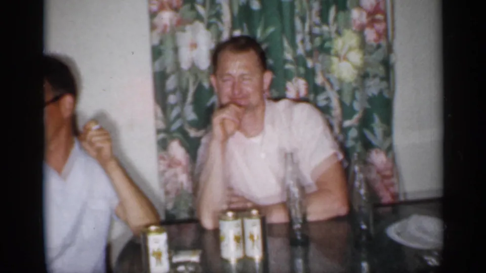 1958 New Jersey home-movie frame showing adult men at a glass table with canned drinks and floral curtains behind them.