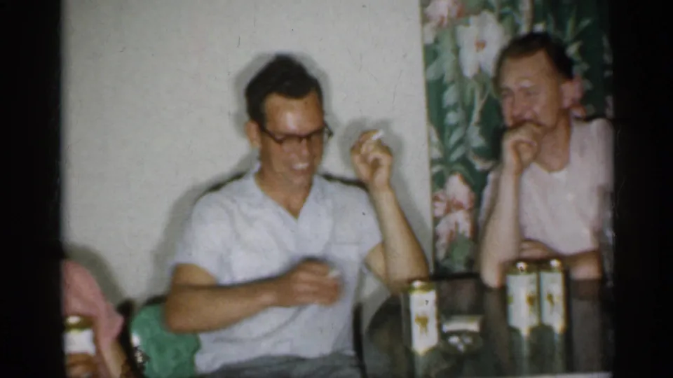 1958 New Jersey frame of a man in glasses smiling with a canned drink as another man sits nearby in a home interior.