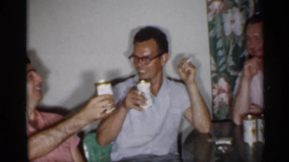 1958 New Jersey home-movie frame showing men around a glass table raising canned beverages in a private room.