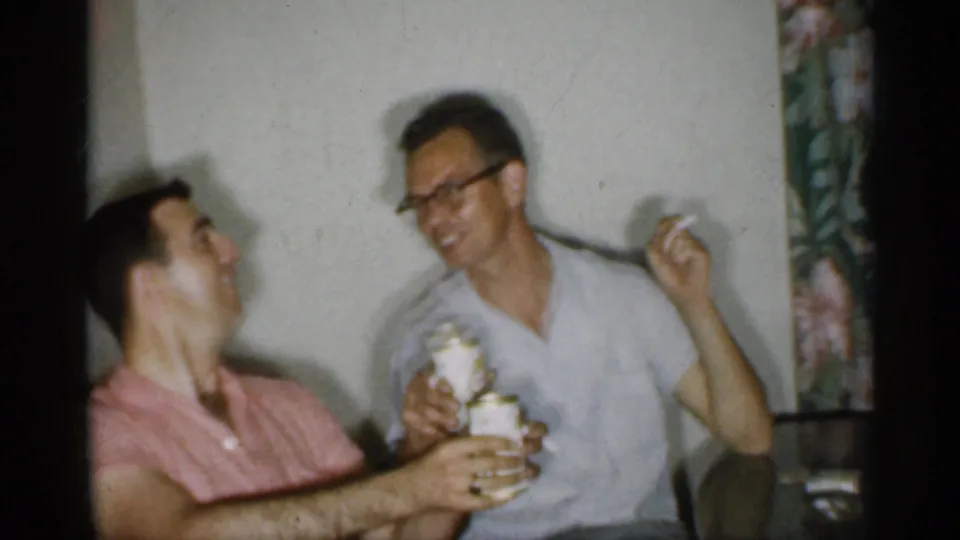 1958 New Jersey frame of two men laughing at a table while holding canned beverages in a home setting.