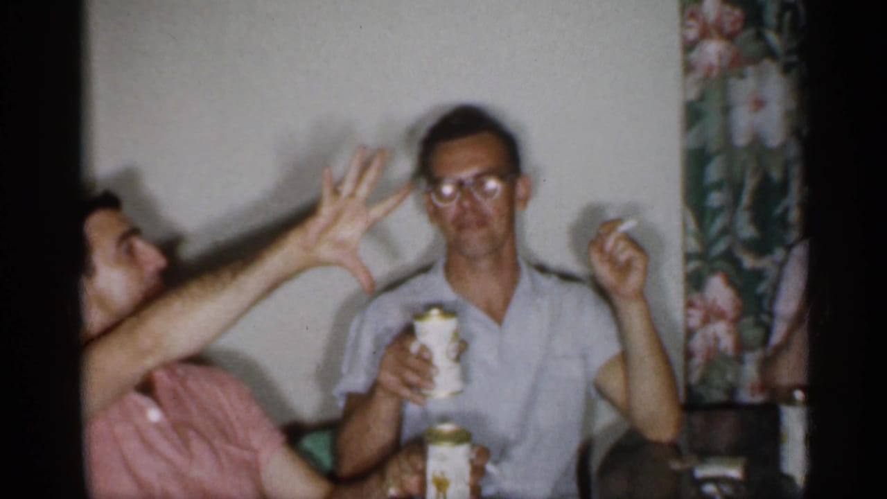 Two adult men seated indoors, one gesturing while the other holds a can and cigarette.