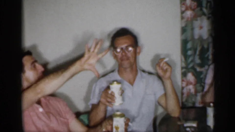 1958 New Jersey home-movie frame of two adult men seated indoors, with cigarettes and beverage cans visible.