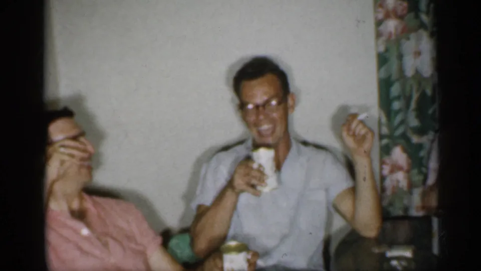1958 New Jersey home-movie frame showing two men relaxing indoors with cigarettes and cans near floral curtains.