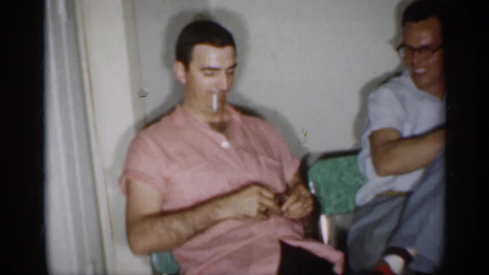 1958 New Jersey home-movie frame of a seated man in a pink shirt holding a cigarette as another man sits nearby.