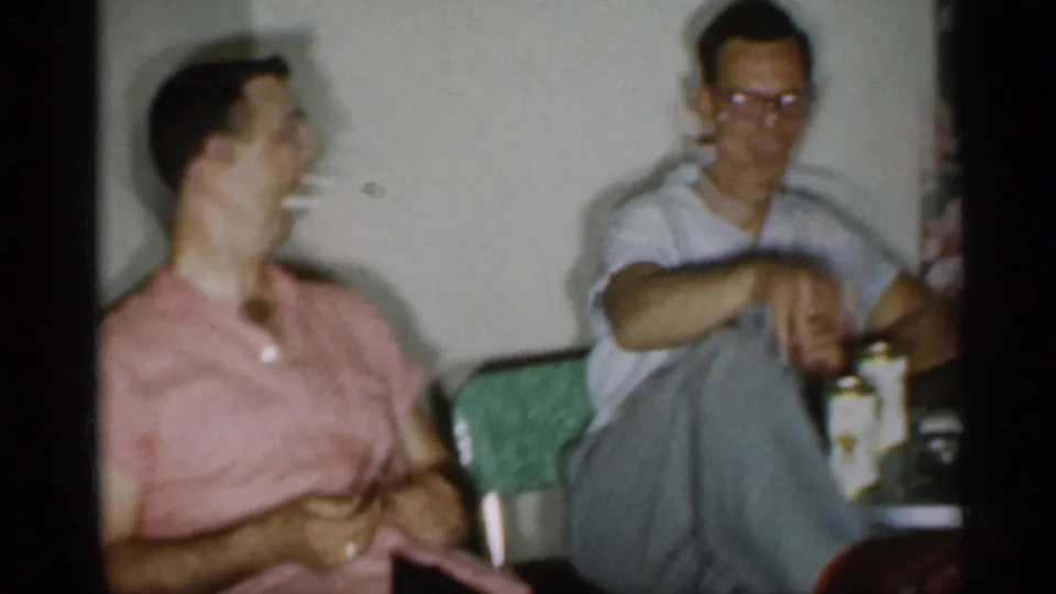 1958 New Jersey home-movie frame of two adult men laughing indoors, with cigarettes and cans visible nearby.