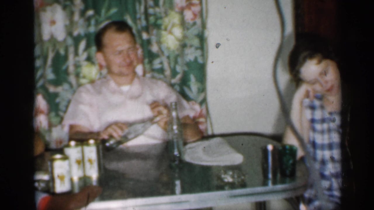 1958 New Jersey home-movie frame of a man seated at a glass-topped kitchen table with drink containers nearby.