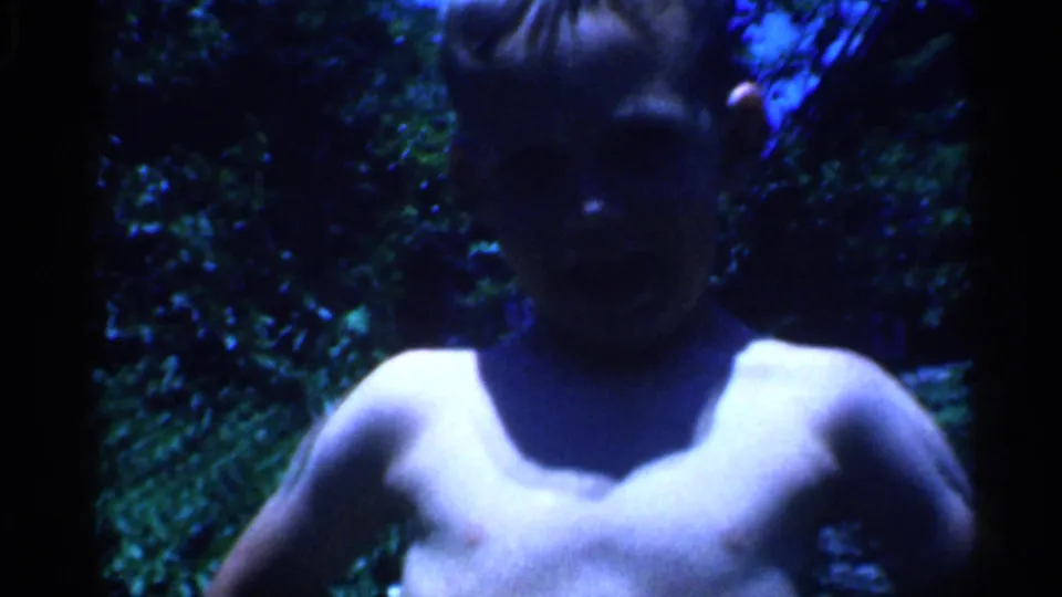 1958 New Jersey 8mm frame showing a shirtless young boy close to the camera, partly shadowed against garden foliage.