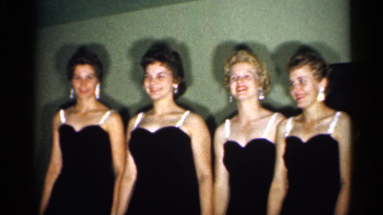 Four women in black dresses stand side by side against a plain wall.