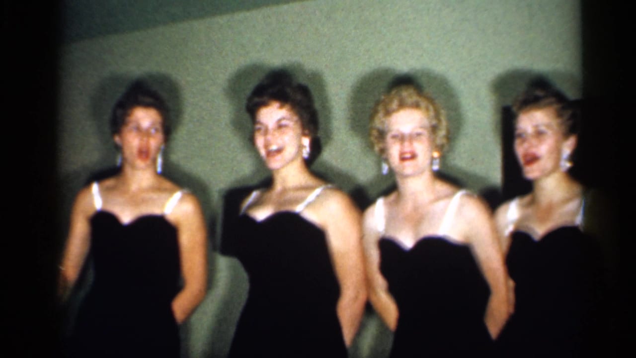 Four young women in dark evening dresses stand against a plain wall.