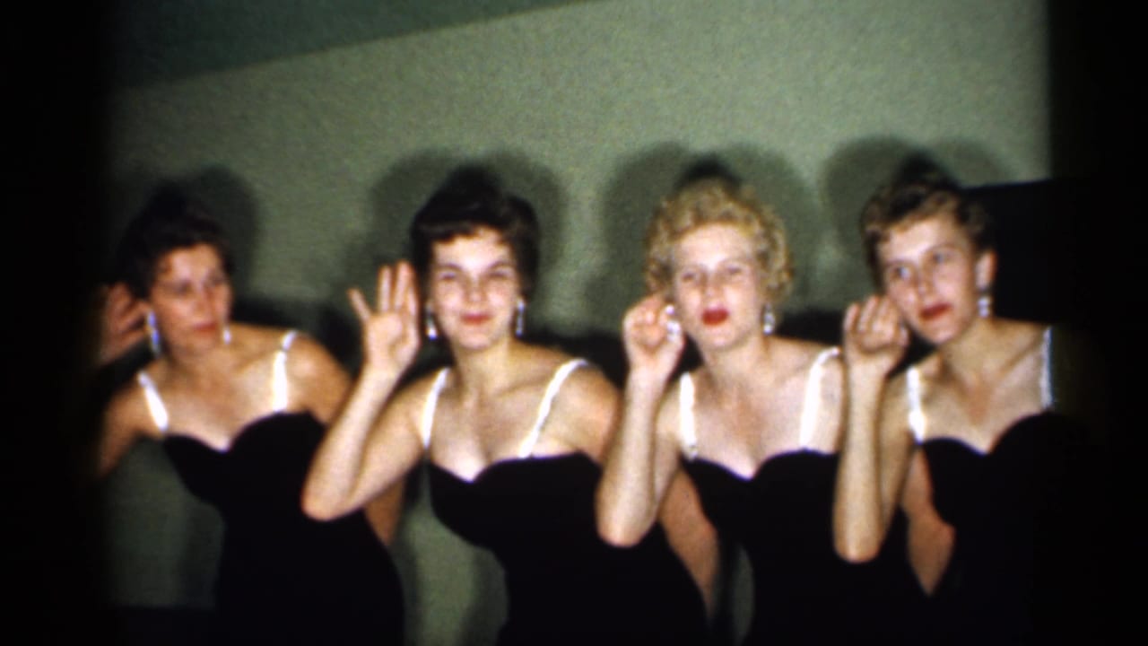 Four formally dressed women hold expressive gestures in a dim 1958 New Jersey indoor home-movie scene.