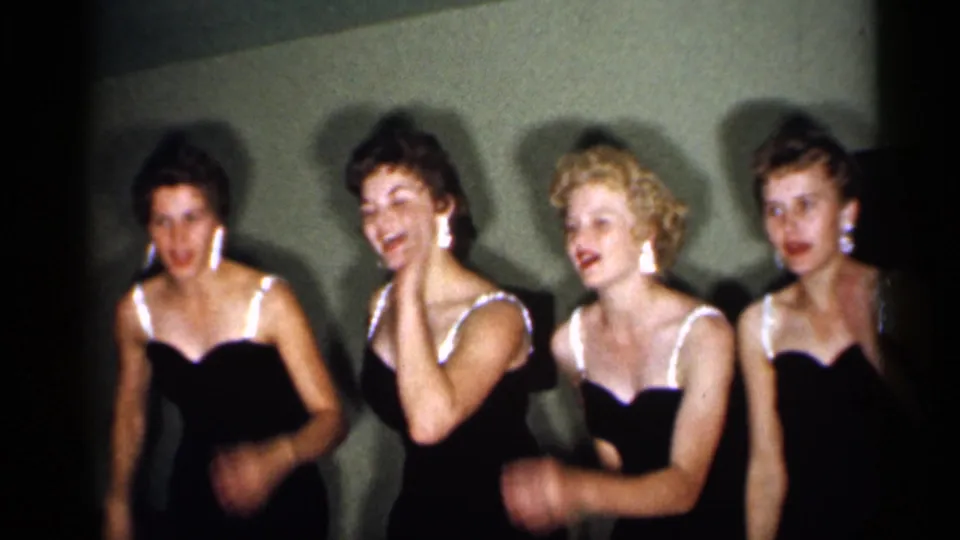 In 1958 New Jersey home-movie footage, four women in matching dark dresses with white straps appear to sing or dance against a plain wall.