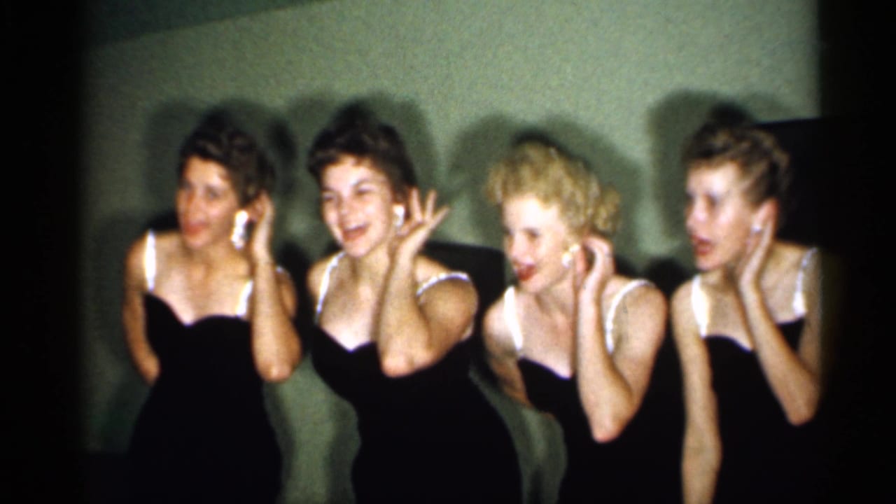 Four adult women in matching black dresses with white trim gesture near their faces while performing for the camera in the 1958 New Jersey clip.