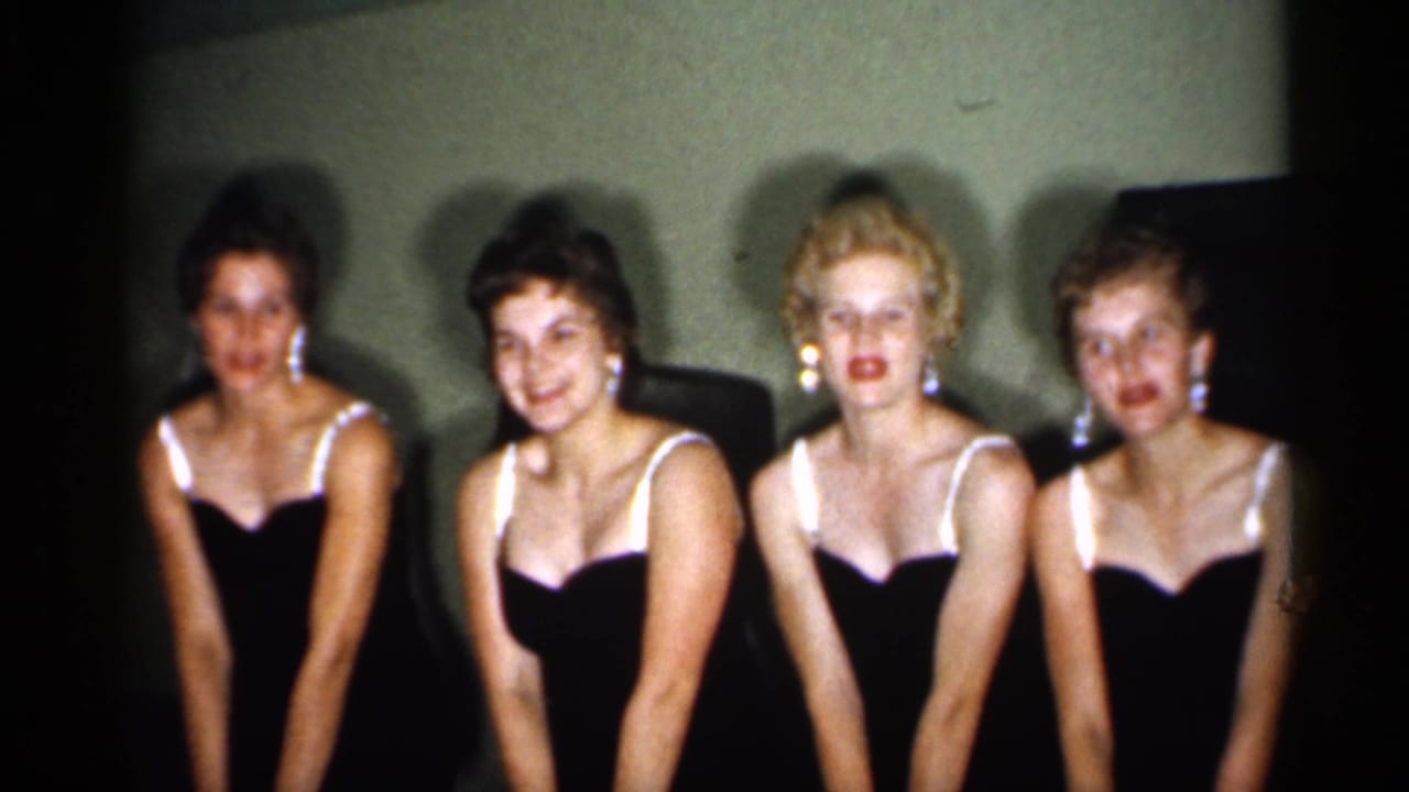 Four adult women in coordinated dark dresses face forward and pose together, with soft 8mm grain and dim interior lighting visible.