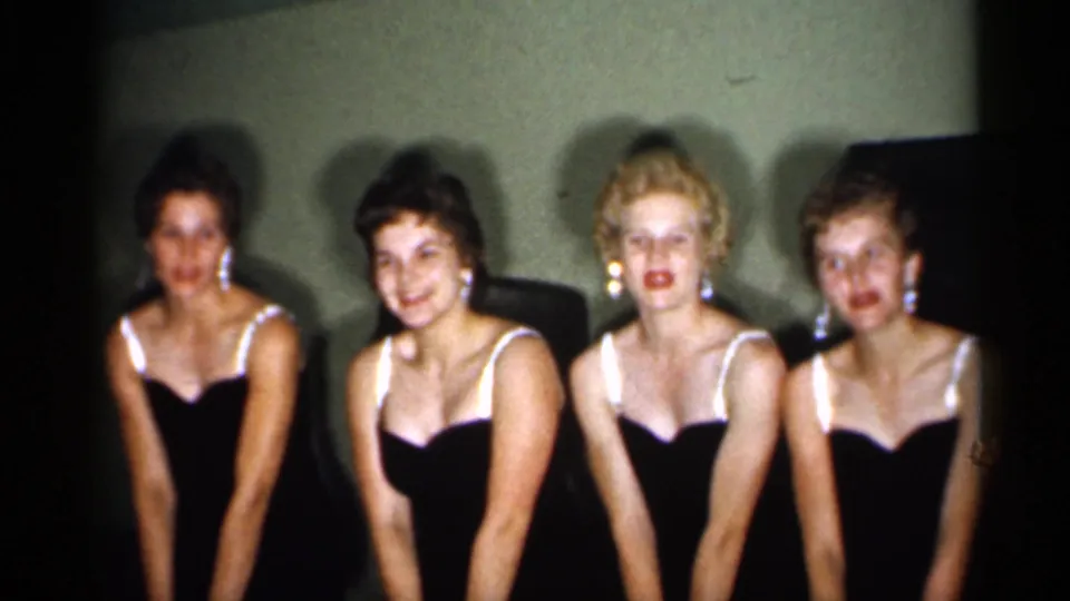 Four adult women in coordinated dark dresses face forward and pose together, with soft 8mm grain and dim interior lighting visible.