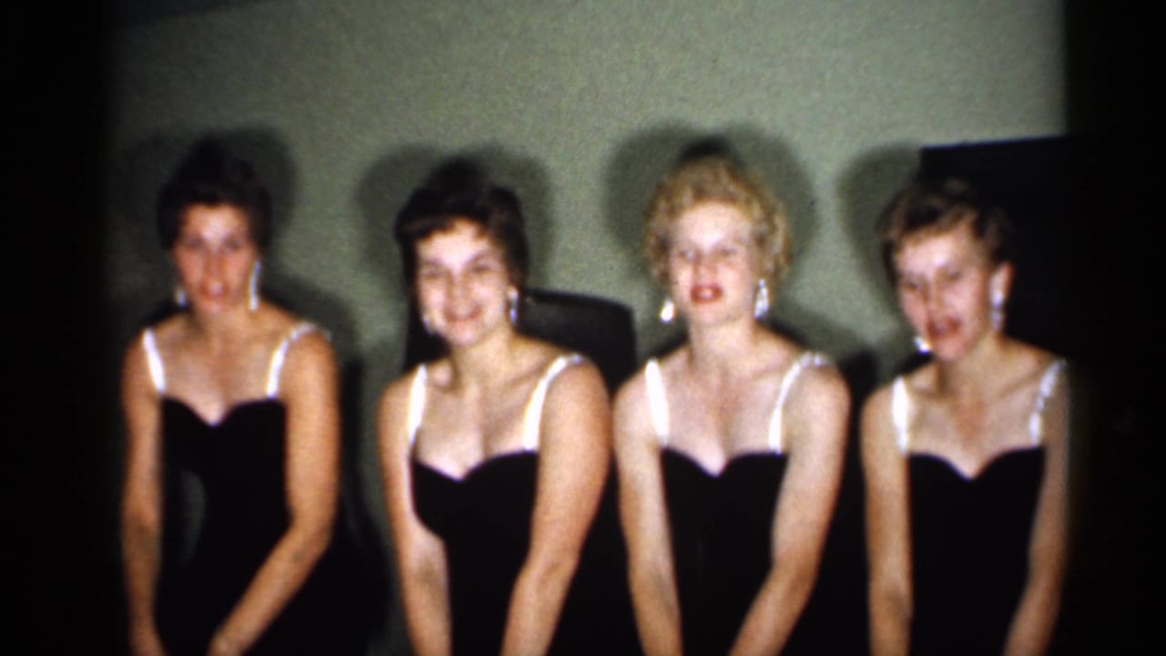 Four young women in dark strap dresses sit or stand together facing the camera.