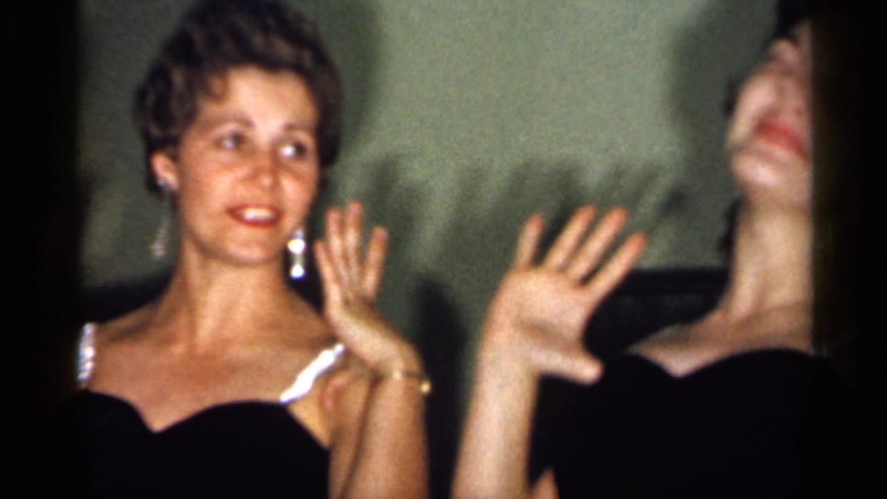 Two young women in dark formal dresses smile and wave indoors in 1958 New Jersey home-movie footage.