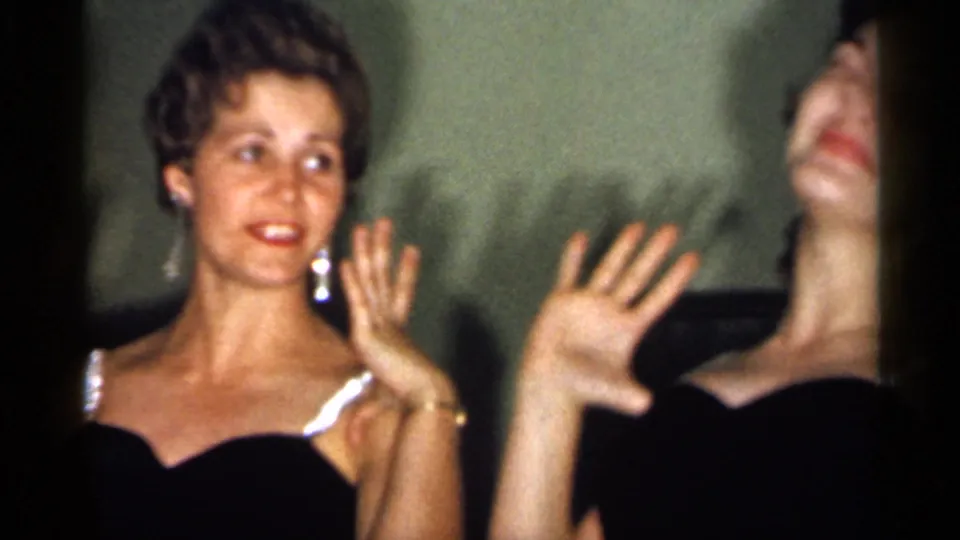 Two young women in dark formal dresses smile and wave indoors in 1958 New Jersey home-movie footage.